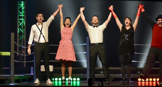 Sarkis Ohanessian, arbitre de la finale France-Québec au Mondial de Catch Impro Professionnel de Genève au Casino Théâtre, lève les bras des vainqueurs. Il est sur scène, dans un ring, entouré de Christian Baumann qui est maître de cérémonie, Igor Potoczny, Selena Hernandez et Roberto Sierra.