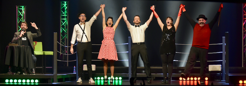 Sarkis Ohanessian, arbitre de la finale France-Québec au Mondial de Catch Impro Professionnel de Genève au Casino Théâtre, lève les bras des vainqueurs. Il est sur scène, dans un ring, entouré de Christian Baumann qui est maître de cérémonie, Igor Potoczny, Selena Hernandez et Roberto Sierra.