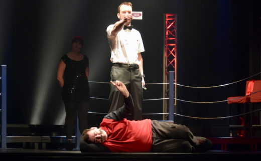 Sarkis Ohanessian, arbitre du Mondial de Catch Impro professionnel est habillé d'un pantalon noir et d'une chemise blanche manches courtes. Il lit le thème de l'improvisation. A ses pieds est couché Roberto Sierra, joueur de l'équipe du Québec.