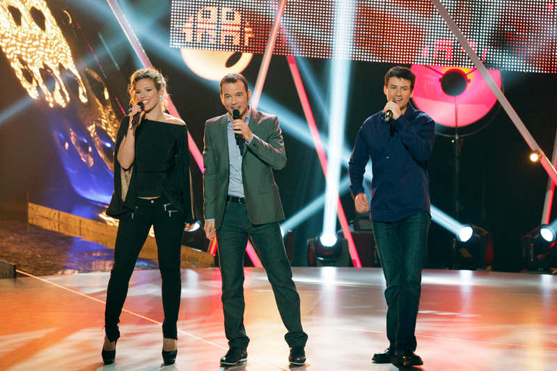 Sarkis Ohanessian, entouré de Lorie Pester et Nicolas Fraissinet, sont sur le plateau d'un Air de Famille, émission de chant produite par la Radio Télévision Suisse. Ils sont tout sourire et chantent l'intro de l'émission. Sarkis est en costume gris, Lorie en jeans noirs et pull noir, Fraissinet en jeans bleu et chemise bleue.