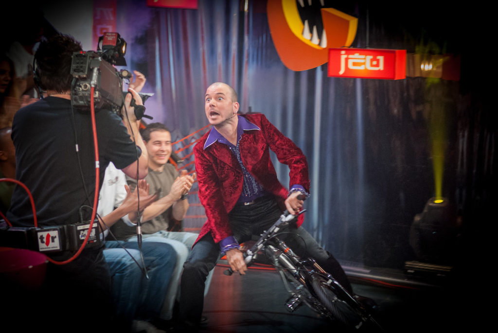 Sarkis Ohanessian arrive à vélo sur le plateau de "C'est mon jeu", comedy game produit par la Radio Télévision Suisse entre 2006 et 2008. Il est plein dérapage et regarde un caméraman. Il est vêtu d'un pantalon noir, une chemise violette à jabot et une veste de costume bordeaux à motifs.