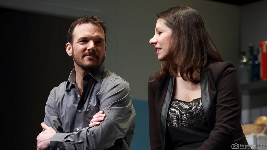 Sarkis Ohanessian, vêtu d'une chemise grise, est sur la scène du Casino Théâtre dans "Cuisine et dépendances" d'Agnès Jaoui et Jean-Pierre Bacri. Il discute avec sa partenaire Estelle.