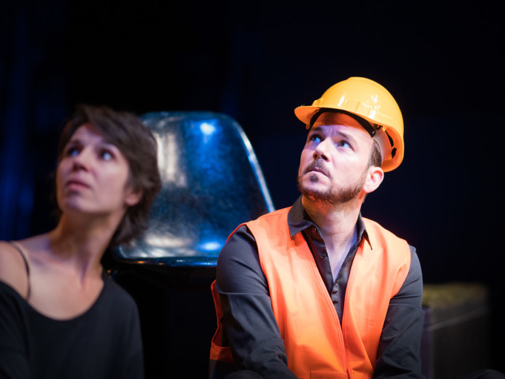 Sarkis Ohanessian est sur la scène du Théâtre Saint-Gervais dans le spectacle improvisé intitulé "dans le décor de...". Il porte un casque de chantier, une chemise noire et un gilet orange. Il regarde vers le haut.