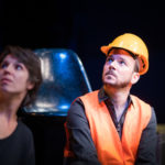 Sarkis Ohanessian est sur la scène du Théâtre Saint-Gervais dans le spectacle improvisé intitulé "dans le décor de...". Il porte un casque de chantier, une chemise noire et un gilet orange. Il regarde vers le haut.