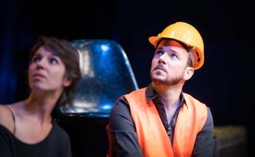 Sarkis Ohanessian est sur la scène du Théâtre Saint-Gervais dans le spectacle improvisé intitulé "dans le décor de...". Il porte un casque de chantier, une chemise noire et un gilet orange. Il regarde vers le haut.