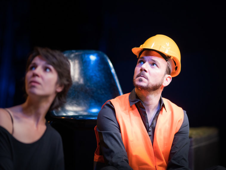 Sarkis Ohanessian est sur la scène du Théâtre Saint-Gervais dans le spectacle improvisé intitulé "dans le décor de...". Il porte un casque de chantier, une chemise noire et un gilet orange. Il regarde vers le haut.