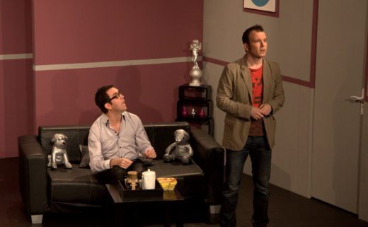 Sarkis Ohanessian est sur la scène du Théâtre Le Caveau dans la pièce "Gros Mensonges" de Luc Chaumar. Il est habillé d'un jeans bleu, d'un t-shirt rouge et d'un veston brun. Il est debout, face public.
