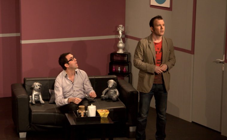 Sarkis Ohanessian est sur la scène du Théâtre Le Caveau dans la pièce "Gros Mensonges" de Luc Chaumar. Il est habillé d'un jeans bleu, d'un t-shirt rouge et d'un veston brun. Il est debout, face public.