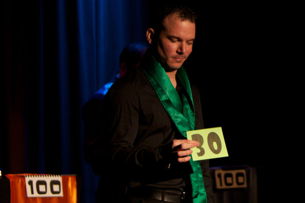 Sarkis Ohanessian est vêtu en chemise noire et pantalon noir. Il porte une écharpe verte satinée autour du cou. Il a dans la main droite une enveloppe avec le nombre 30 marqué dessus. Il joue le spectacle d'improvisation intitulé "le Grand Jeu".