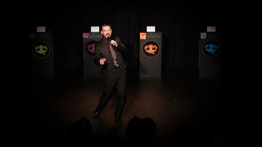 Sarkis Ohanessian présente le spectacle d'importation intitulé "le Grand jeu". Il est vêtu d'un pantalon noir, une chemise noire et une cravate bariolée.
