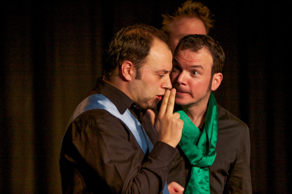 Sarkis Ohanessian est en train de jouer le spectacle d'improvisation intitulé "le Grand Jeu". Il porte une chemise noire et une écharpe de verte satinée.