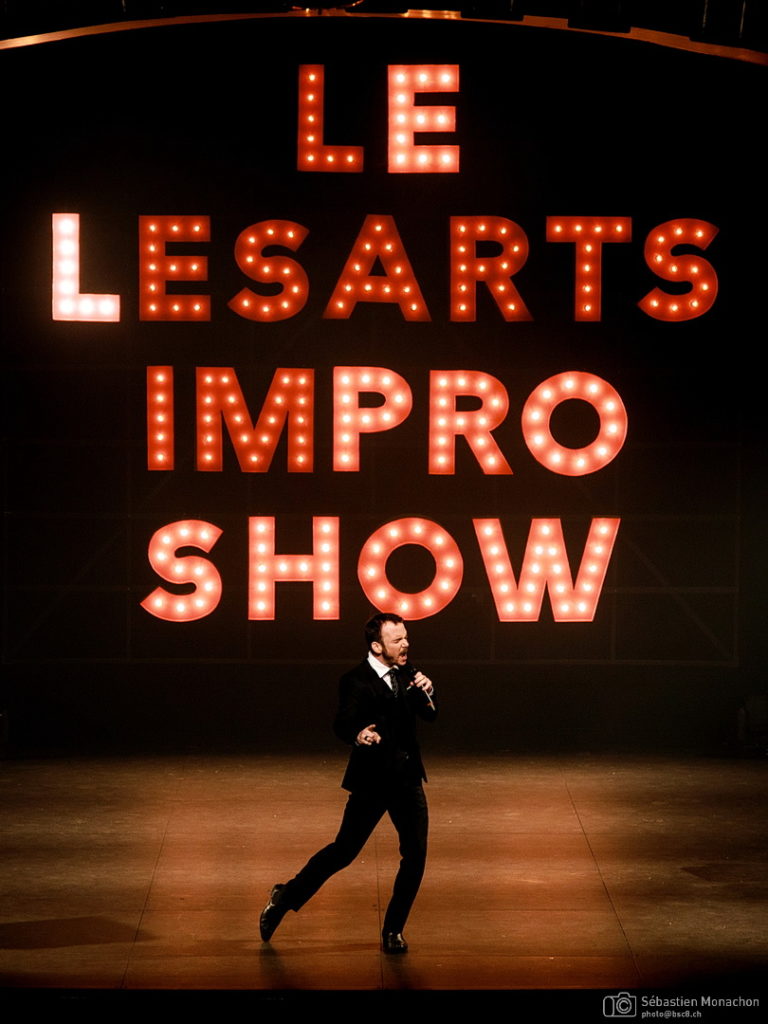 Sarkis Ohanessian, très classe, vêtu d'un costume noir, d'une chemise blanche et d'une cravate noire, est sur la scène du Casino Théâtre de Genève et met le feu au public. Derrière lui, en fond de scène, des panneaux lumineux écrivent "Le lésants Impro Show"