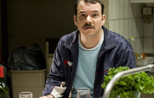 Sarkis Ohanessian, sur le tournage d'Opération Casablanca, joue Gilbert. Il porte une moustache, un pull bleu clair et une veste de training bleu foncé. Il est assis à table avec un verre de rouge devant lui et regard l'objectif d'un air un peu hébété.