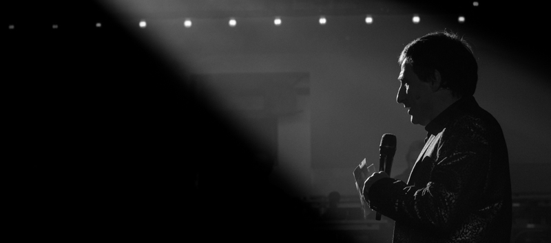 Sur une photo en noir et blanc, Benjamin Cuche, présentateur en alternance avec Sarkis Ohanessian de la Grande Revue Improvisée de Servion, se trouve éclairé de côté par un spot. Il se trouve dans le public, micro à la main.
