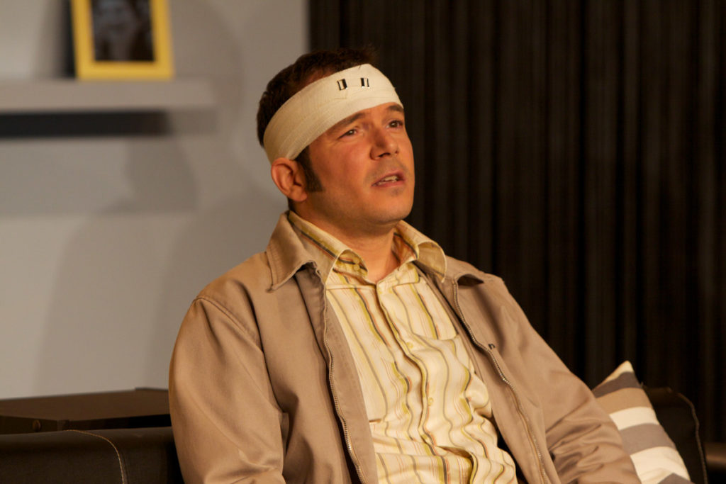 Sarkis Ohanessian est sur la scène du théâtre Le Caveau. Il est en gros plan, assis sur un canapé. Il porte un bandage à la tête et à l'air d'avoir mal. Il est habillé d'une chemise jaune rayée et d'une veste grise.