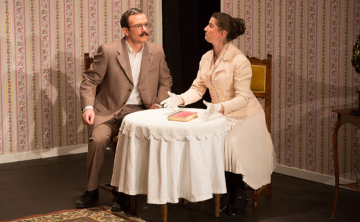 Sarkis Ohanessian est sur la scène du Théâtre Le Caveau dans "Thé à la menthe ou t'es citron" de Patrick Haudecoeur. Il est habille d'un costume gris et porte des lunettes et une moustache. Il est assis à table avec Julie Despriet, elle est en train de lui expliquer quelque chose. La table est recouverte d'une nappe blanche et un livre est posé dessus.