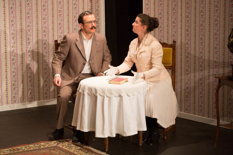 Sarkis Ohanessian est sur la scène du Théâtre Le Caveau dans "Thé à la menthe ou t'es citron" de Patrick Haudecoeur. Il est habille d'un costume gris et porte des lunettes et une moustache. Il est assis à table avec Julie Despriet, elle est en train de lui expliquer quelque chose. La table est recouverte d'une nappe blanche et un livre est posé dessus.