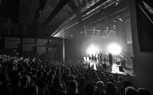 Saluts finaux de la Coupe du Monde d'impro catch au Théâtre du Passage à Neuchâtel. Plan large de la scène avec le public. L'arbitre lève les mains des vainqueur, des feux d'artifice éclairent la scène.