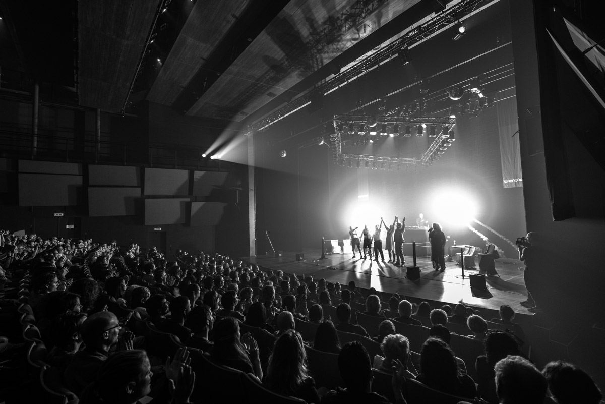 Saluts finaux de la Coupe du Monde d'impro catch au Théâtre du Passage à Neuchâtel. Plan large de la scène avec le public. L'arbitre lève les mains des vainqueur, des feux d'artifice éclairent la scène.