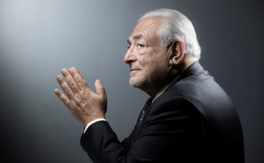 Dominique Strauss-Kahn est de profil, les mains jointes. Il porte un costume noir.