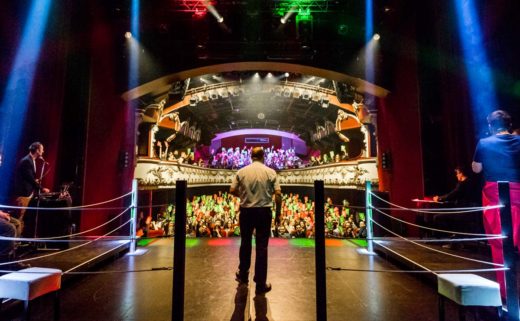 La scène du Casino Théâtre vue de dos lors du Mondial de Catch Impro Professionnel de Genève. L'arbitre, François Briard, est habillé d'un pantalon noir et d'une chemise blanche. Au fond, on voit les spectateurs en train de voter.