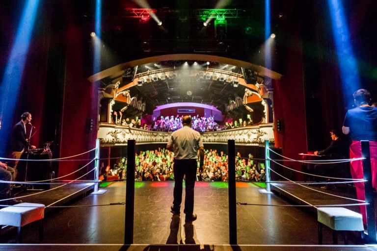 La scène du Casino Théâtre vue de dos lors du Mondial de Catch Impro Professionnel de Genève. L'arbitre, François Briard, est habillé d'un pantalon noir et d'une chemise blanche. Au fond, on voit les spectateurs en train de voter.