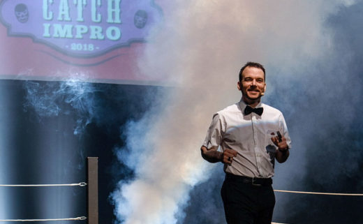 Sarkis Ohanessian, arbitre du Mondial de Catch Impro Professionnel de Genève est en train de s'avancer sur le ring poser sur la scène. Il est vêtu d'un pantalon noir, d'une chemise blanche avec un noeud papillon noir. Il est entouré de fumée et au fond derrière lui est projeté l'affiche du Mondial.