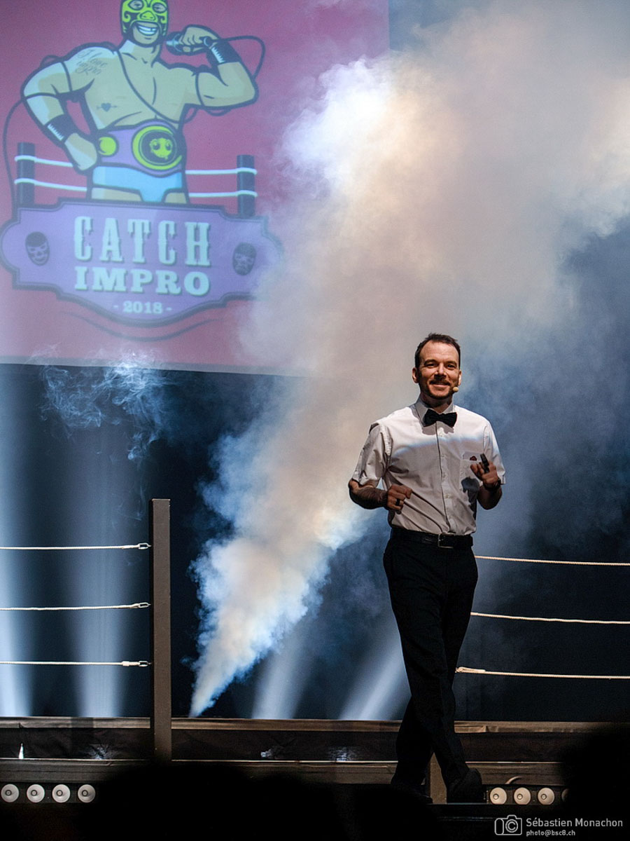 Sarkis Ohanessian, arbitre du Mondial de Catch Impro Professionnel de Genève est en train de s'avancer sur le ring poser sur la scène. Il est vêtu d'un pantalon noir, d'une chemise blanche avec un noeud papillon noir. Il est entouré de fumée et au fond derrière lui est projeté l'affiche du Mondial.