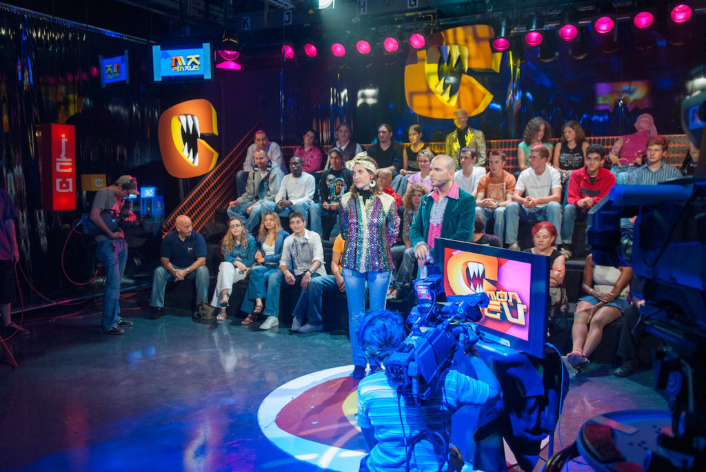Sarkis Ohanessian, vêtu d'une chemise rose à jabot et d'une veste de costume verte, présente "C mon jeu"; un comedy game de la Radio Télévision Suisse. Il a à côté de lui une candidate habillée d'une chemise à paillettes. On voit le public de l'émission derrière eux.