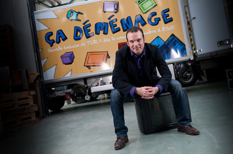 Sarkis Ohanessian est assis sur une télévision devant le camion de l'émission "Ça déménage" qu'il présente pour la Radio Télévision Suisse. il Est vêtu d'un jean bleu, une chemise violette et une veste noire.