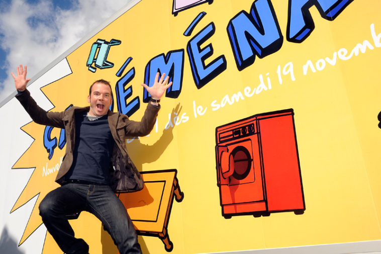 Sarkis Ohanessian saut d'un camion sur lequel est écrit "Ça déménage", titre de l'émission qu'il anime pour la Radio Télévision Suisse. Il porte un jean noir, un pull bleu foncé et une veste brune.