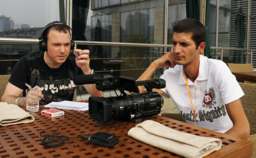 Sarkis Ohanessian, lors de championnats du monde de magie à Pékin, est attablé, un casque sur les oreilles. Il tient un stylo à la main, sur la table sont posés un jeu de cartes, une caméra, un bloc de papier et un micro. A côté de lui se trouve le réalisateur et caméraman Michael Dirani.