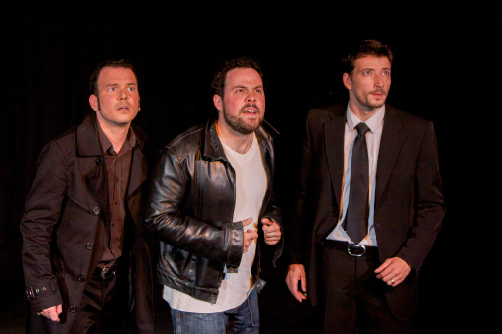 Sarkis Ohanessian, avec Alexandre de Marco et Jérôme Ricca sur la scène du Théâtre le Caveau dans la pièce "Les Rogers" de Jean Marc Dalpé, Robert Bellefeuille et Robert Marinier. Les trois comédiens regardent vers le haut. Sarkis est habillé d'un jean, chemise et manteau.
