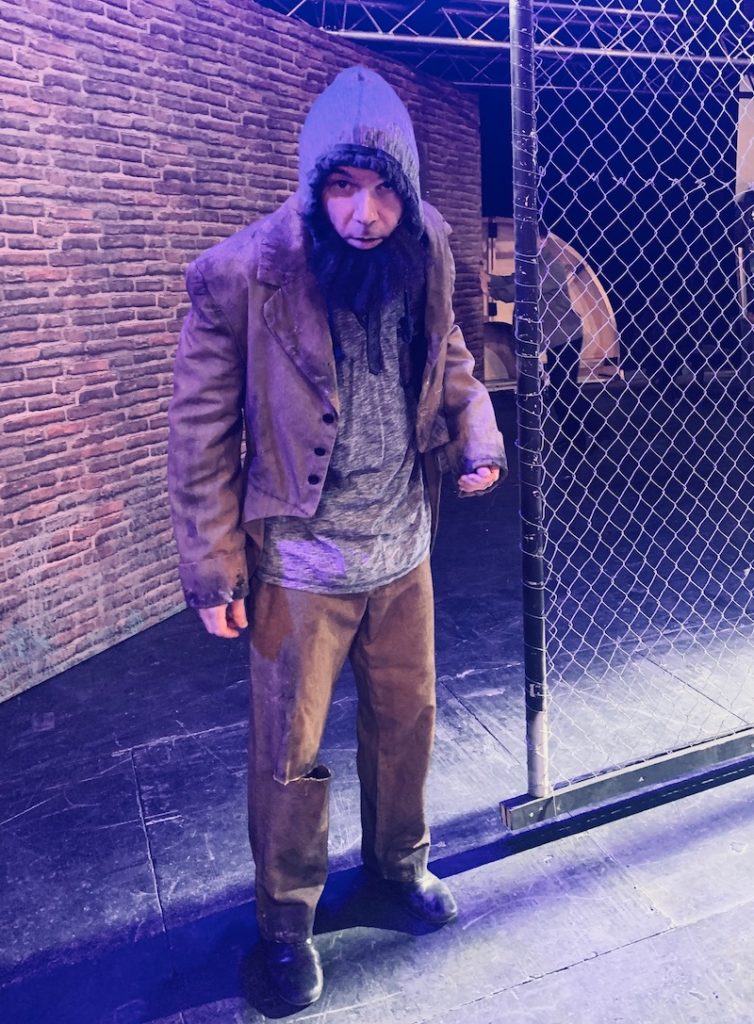 Sarkis Ohanessian est habillé en clochard pour le spectacle "Sister Act" au théâtre Barnabé de Servion. Il porte une fausse barbe noire fournie, un pantalon vert déchiré au niveau du genou, un sweat à capuche gris et un manteau vert.