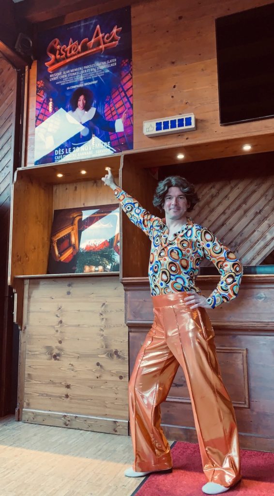 Sarkis Ohanessian est habillé en danseur disco pour le spectacle "Sister Act" au théâtre Barnabé de Servion. Il porte une chemise à motifs ronds et un pantalon orange pattes d'éléphant orange brillant. Il a une perruque sur la tête.