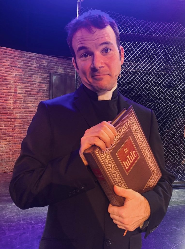 Sarkis Ohanessian est habillé en prêtre pour le spectacle "Sister Act" au théâtre Barnabé de Servion. Il porte un costume noir et tient une grosse bible dans les mains.