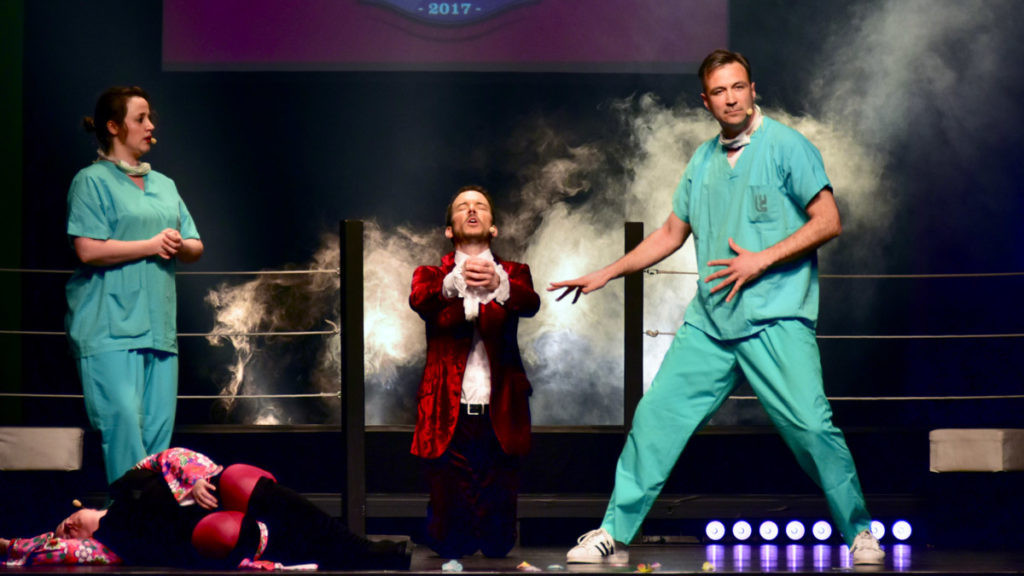 Sarkis Ohanessian est sur la scène du Casino Théâtre pour la rencontre Suisse-Belgique du Mondial de Catch Impro Professionnel à Genève. Il est vêtu d'un pantalon violet, d'une chemise blanche à jabot et d'une veste rouge à motifs. Il est entouré de Gilles Delveux, Amandine Vandormael et Julie Despriet qui est allongée au sol.