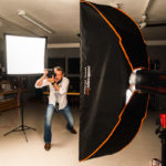 Sarkis Ohanessian est en plein séance de photographie en studio. Il tient son appareil verticalement et est entouré de flashes. Il porte un jean bleu et une chemise blanche et a un posemètre autour du cou.