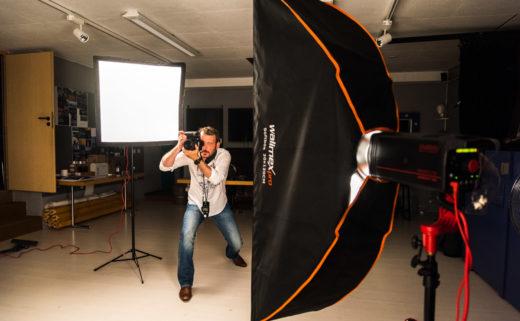 Sarkis Ohanessian est en plein séance de photographie en studio. Il tient son appareil verticalement et est entouré de flashes. Il porte un jean bleu et une chemise blanche et a un posemètre autour du cou.