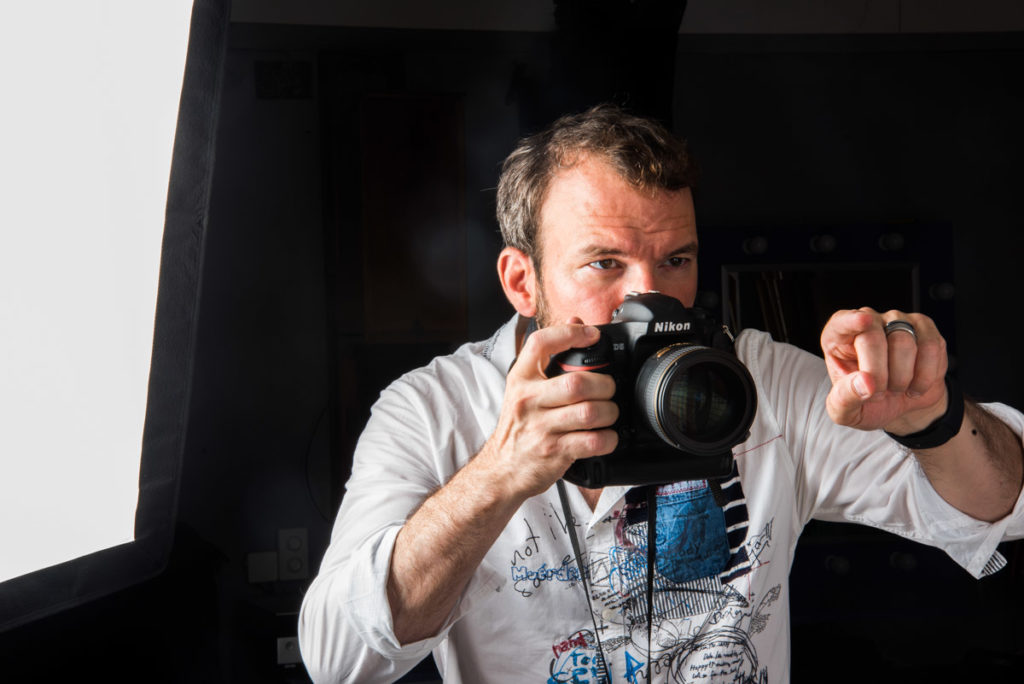 Sarkis Ohanessian est en plein séance de photographie en studio. Il tient son appareil de photo dans la main droit, pointe son doigt gauche vers le modèle. Il porte une chemise blanche.