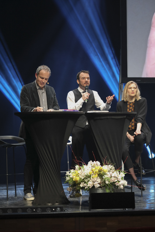 Sarkis Ohanessian, en chemise blanche et gilet noir, présente anime la partie témoignages des Promotions Citoyennes de la Ville de Genève. Il est entouré de Salomé Joly et Zep, dessinateur de Titeuf.