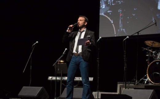 Sarkis Ohanessian est en train de présenter les Promotions Citoyennes de la Ville de Genève sur la scène du Théâtre du Léman. Il porte un jean bleu, une chemise blanche, une veste de costume noire légèrement zébrée et une cravate noire rayée de lignes blanches. Il tient dans sa main droite son micro et dans sa main gauche des fiches.