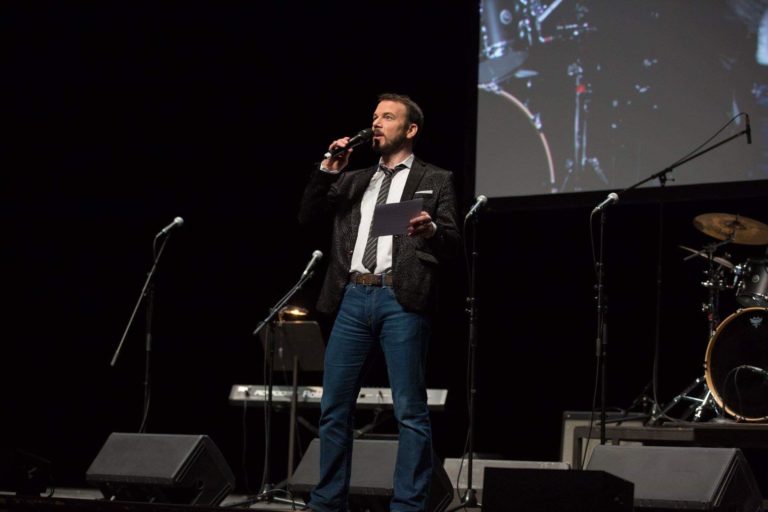 Sarkis Ohanessian est en train de présenter les Promotions Citoyennes de la Ville de Genève sur la scène du Théâtre du Léman. Il porte un jean bleu, une chemise blanche, une veste de costume noire légèrement zébrée et une cravate noire rayée de lignes blanches. Il tient dans sa main droite son micro et dans sa main gauche des fiches.