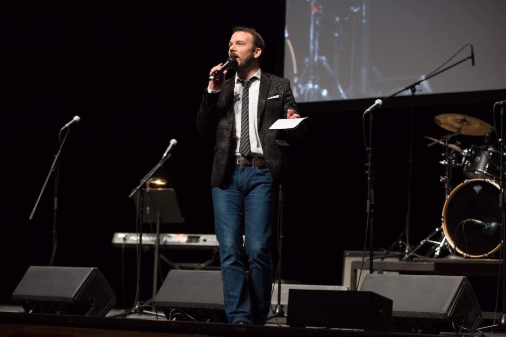 Sarkis Ohanessian est en train de présenter les Promotions Citoyennes de la Ville de Genève sur la scène du Théâtre du Léman. Il porte un jean bleu, une chemise blanche, une veste de costume noire légèrement zébrée et une cravate noire rayée de lignes blanches. Il tient dans sa main droite son micro et dans sa main gauche des fiches.