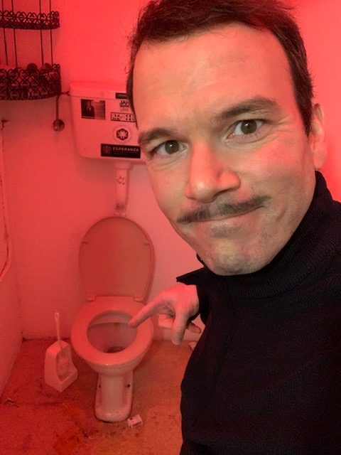 Sarkis Ohanessian, dans le rôle de Boris, porte une moustache. Il montre du doigt la cuvette des toilettes.