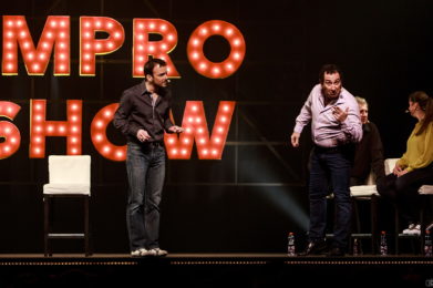 Photo couleur de Sarkis Ohanessian en plein spectacle d'improvisation théâtrale. Il est vêtu d'un jeans bleu et d'une chemise brune. Il est accompagné de Fausto Borghini, Julie Despriet et Jean-Claude Dubiez. Des tabourets blancs sont sur scène et au fond un panneau lumineux indique "Impro Show".