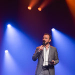 Sarkis Ohanessian, costume gris et chemise blanche, est sur une scène et des projecteurs de couleurs sont dirigés sur lui.