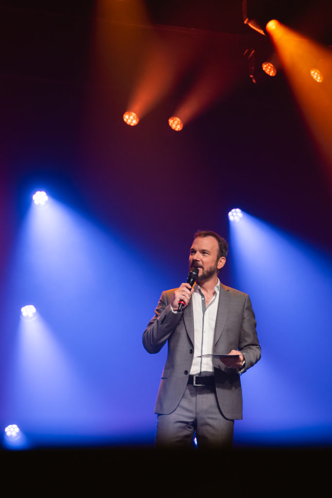 Sarkis Ohanessian, costume gris et chemise blanche, est sur une scène et des projecteurs de couleurs sont dirigés sur lui.