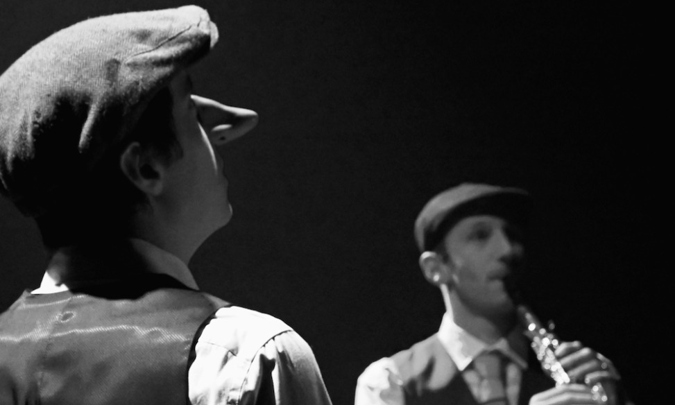 Image en noir et blanc montrant deux comédiens, un de dos avec un bey imposant (Cyrano) et un autre de face jouant de la clarinette